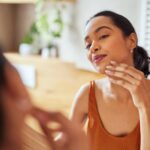 mulher se olhando no espelho e passando a mão no rosto para ilustrar o conteúdo sobre tipos de pele