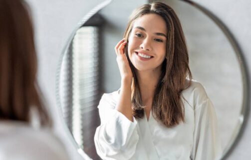 mulher na frente do espelho passando a mão no rosto para ilustrar o conteúdo sobre vitamina C