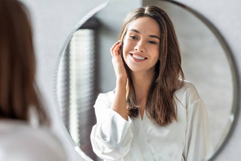 mulher na frente do espelho passando a mão no rosto para ilustrar o conteúdo sobre vitamina C
